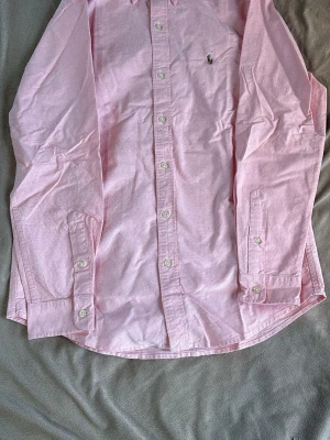Rosa skjorta från Ralph Lauren - Klassisk rosa skjorta från Ralph Lauren med broderad logga på bröstet. Skjortan har lång ärm, knappar framtill och är tillverkad i bomull. Perfekt för en stilren och avslappnad look.