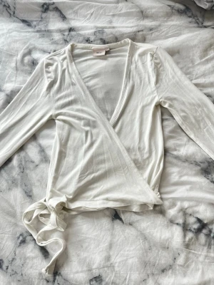 Wrap tie topp - Säljer en vit wrap tie topp från Nelly med långa ärmar och knytband i sidan. Toppen har v-ringning och är tillverkad i mjukt, tunt material som känns skönt mot huden. Perfekt att styla med jeans eller kjol.