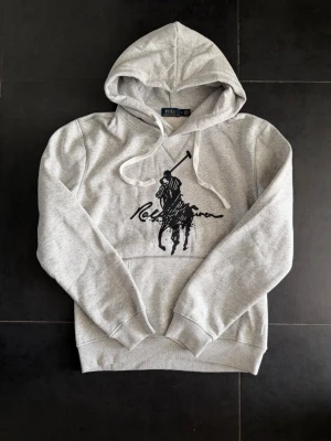 Grå hoodie från Ralph Lauren - Snygg grå hoodie från Ralph Lauren med stor svart polospelare och logga på bröstet. Klassisk känguruficka framtill, ribbade muddar och justerbar huva med vita snören. Tillverkad i mjukt sweatshirtmaterial som är perfekt för chill dagar. Storlek M men sitter mer åt S hållet ✅