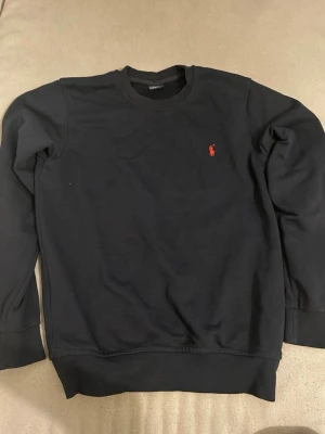 Svart sweatshirt från Ralph Lauren - Klassisk svart sweatshirt från Ralph Lauren med rund halsringning och den ikoniska röda logotypen broderad på bröstet. Tröjan har ribbade muddar vid ärmslut och nederkant, perfekt för en clean och stilren look.