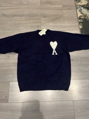 Mörkblå stickad tröja från Ami Paris - Mörkblå stickad tröja från Ami Paris med rund halsringning och ribbade muddar. På bröstet finns ett vitt hjärta och bokstaven A, vilket är typiskt för märket. Perfekt för en stilren och trendig look. Passar mer som L
