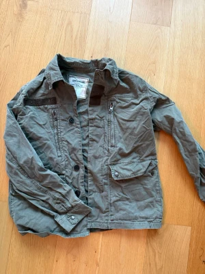 Grön jacka från Zadig & Voltaire - Snygg grön jacka från Zadig & Voltaire med flera fickor, dragkedjor och knappar framtill. Jackan har klassisk krage och är tillverkad i bomull. Jackan är ganska liten i storleken 