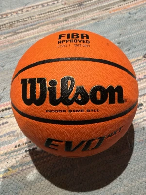 Wilson EVO NXT basketboll storlek 7 - Wilson EVO NXT basketboll i orange med svarta detaljer. FIBA-godkänd för inomhusspel, tillverkad i slitstarkt syntetmaterial. Perfekt för matcher och träning, officiell storlek och vikt. Tydlig logga och greppvänlig yta. Det står LG på bollen men går lätt och ta bort med lite handsprit.