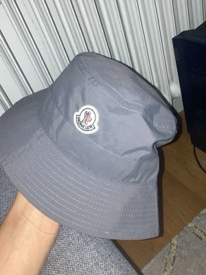 Grå bucket hat från Moncler - Snygg grå bucket hat från Moncler med klassisk logotyp framtill. Hatten har bred skärm och justerbar dragsko baktill för perfekt passform. Tillverkad i ett lätt och slitstarkt material som passar året runt. Perfekt accessoar för en trendig streetwear-look.