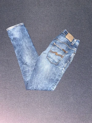 Blå jeans från Nudie Jeans - Säljer ett par klassiska blå jeans från Nudie Jeans med snygg slitning och tvättad look. Modellen har fem fickor, normal midja och smal passform. Bakfickan har Nudie Jeans ikoniska broderi. Perfekta för dig som gillar en avslappnad men stilren vibe. Storlek W32 L36