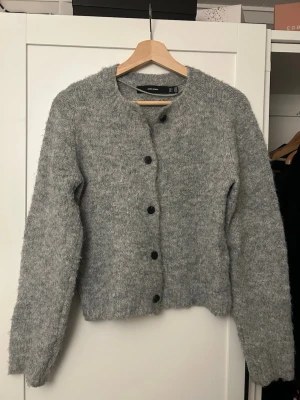 Grå fluffig kofta från Vero Moda - Mysig grå kofta från Vero Moda med fluffig textur och rund halsringning. Koftan har svarta knappar framtill och långa ärmar💞