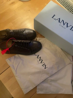Lanvin med ormmönster - Helt nya snygga svarta sneakers från Lanvin med glansigt ormmönstrat material och svarta snören. Låg modell med klassisk siluett och metalliska öljetter. Perfekta för dig som vill sticka ut med en unik textur och modern look. Nypris 11 999