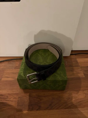 Svart Gucci skärp med orm-motiv - Säljer ett svart skärp från Gucci med ikoniskt GG-mönster och en rödvit orm längs hela bandet. Skärpet har silverfärgat spänne och är gjort i skinn. Perfekt för dig som vill ha en statement-accessoar från ett lyxigt märke.