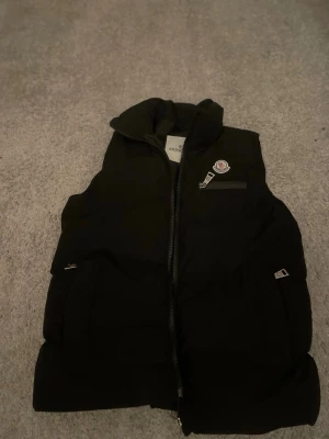 Svart dunväst från Moncler - Snygg svart dunväst från Moncler med hög krage och dragkedja framtill. Västen har två sidofickor med dragkedja och en bröstficka med Moncler-logga. Insidan har ett färgglatt tygmärke. Perfekt för lager-på-lager och streetwear.helt äkta 