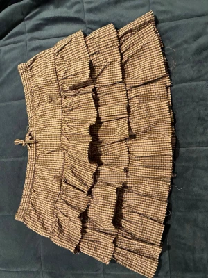 Brun- och svartrutig volangkjol - Kort volangkjol i brunt och svart med rutigt mönster. Kjolen har flera lager volanger och ett elastiskt midjeband med liten knytning. Perfekt för dig som gillar en söt och avslappnad stil.