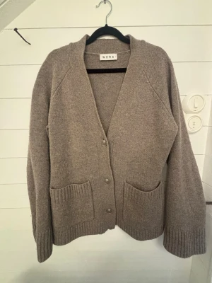 Beige kofta från Wera med fickor - Mysig beige ull kofta från Wera med v-ringning, knäppning framtill och två stora fickor. Koftan har ribbade muddar vid ärmslut och nederkant, samt en avslappnad passform. 