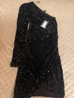 Svart paljettklänning omlott - Snygg svart kortklänning täckt med glittriga paljetter och omlottdetalj framtill. Klänningen har one shoulder design och en figurnära passform som ger en festlig look. Perfekt för dig som vill sticka ut på nästa event. Klänning är sydd lite vid sidan då det va stor i storlek, kan enkelt sprättas upp.💕