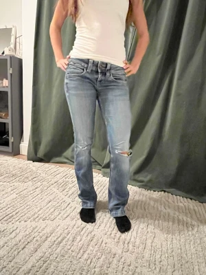 Blå slitna bootcut jeans  - Snygga blå bootcut jeans och slitningar på knäna. Jeansen har klassisk femficksdesign och låg midja. Perfekta för en avslappnad och trendig look.