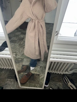 Beige lång teddy-kappa med knytskärp - Säljer en beige lång teddy-kappa med knytskärp i midjan och bred krage. Kappan är gjord i ett mjukt och fluffigt material som håller dig varm under kalla dagar. Perfekt för dig som gillar en avslappnad och trendig look.