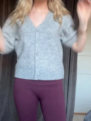 Grå kortärmad kofta med knappar - Snygg grå kofta med korta puffärmar och v-ringning. Koftan har ribbade muddar och knäpps framtill med matchande knappar. Perfekt att slänga över en topp för en chill look. Materialet ser ut att vara mjukt och stickat. Kollar intresse!