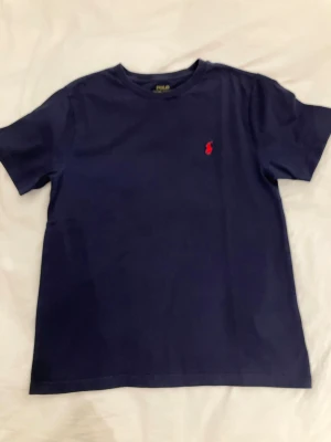 Mörkblå t-shirt från Polo Ralph Lauren i mycket bra skick storlek 152 - En stilren mörkblå t-shirt från Polo Ralph Lauren med klassisk passform och röd broderad logga på bröstet. T-shirten har rund halsringning och är tillverkad i mjuk bomull, perfekt för en avslappnad look. Storlek 152 i mycket bra skick. 