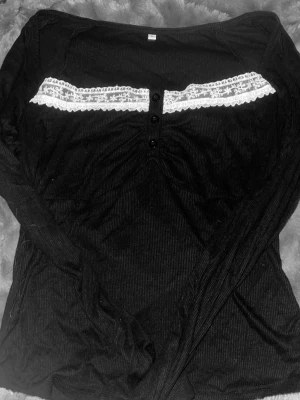 Svart långärmad topp med spets - Svart ribbad långärmad topp med dekorativ vit spets och knappar framtill. Toppen har en feminin och trendig look med smickrande passform och är perfekt att styla till jeans eller kjol.