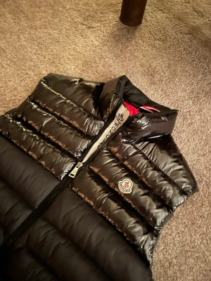 Svart dunväst från Moncler - Snygg svart dunväst från Moncler med blank finish och röd insida. Västen har ståkrage, två fickor med tryckknappar och dragkedja framtill. Klassisk Moncler-logga på bröstet. Alltså en väldigt bra/snygg väst inför sommaren,våren,hösten 