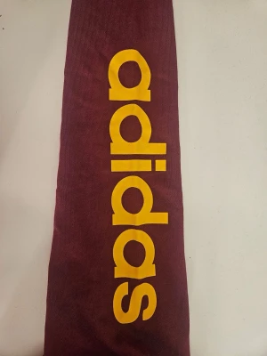 Vinröd hoodie från Adidas - Snygg vinröd hoodie från Adidas med stor gul logga längs ena ärmen. Klassisk huva och ribbade muddar. Perfekt för dig som vill ha en sportig och trendig look. Tillverk excoolt bomullsmaterial som känns skönt mot huden pam vardagen. Helt ny!