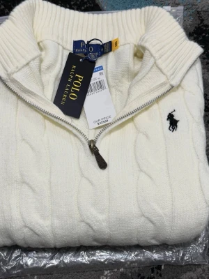 Vit kabelstickad tröja Polo Ralph Lauren - Kabelstickad vit tröja från Polo Ralph Lauren med halv dragkedja och hög krage. Tröjan har det klassiska broderade logotypen i marinblått på bröstet och ribbstickade muddar. Perfekt för dig som gillar stilren och tidlös design.