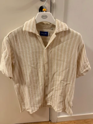 Beige randig skjorta från Jack & Jones - Kortärmad skjorta från Jack & Jones i beige och vitt med diskret randigt mönster. Skjortan har klassisk krage, knappar framtill och är tillverkad i ett luftigt bomullstyg som passar perfekt till varmare dagar.