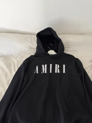 Svart hoodie från Amiri - Svart hoodie från Amiri med stor vit logga framtill. Klassisk modell med huva och känguruficka. Perfekt för en avslappnad streetwear-look. Tillverkad i mjukt material som känns skönt mot huden.