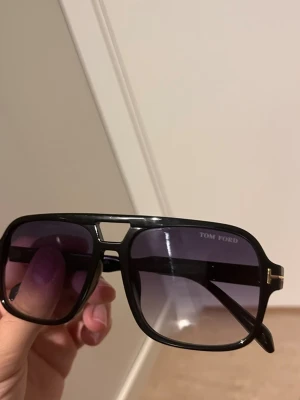 Svarta Tom Ford pilotbågar solglasögon - Snygga svarta solglasögon från Tom Ford med klassisk pilotform och mörka tonade glas. Bågarna har gulddetaljer vid sidorna och en cool brygga över näsan. Perfekta för dig som vill ha en stilren och trendig accessoar.