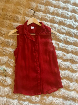 Röd ärmlös blus från Vero Moda - Snygg röd ärmlös blus från Vero Moda i storlek XS. Blusen har krage, knappar framtill och ett lätt, luftigt tyg med struktur. Perfekt till sommaren och enkel att styla med jeans eller kjol.