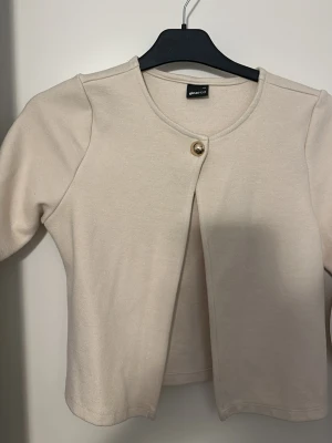 Beige kofta från Gina Tricot - Snygg beige kofta från Gina Tricot med rund halsringning och en dekorativ knapp framtill. Koftan har en enkel och stilren design med långa ärmar och mjukt material. Perfekt att slänga över en topp för en clean look.
