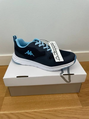 Marinblå sneakers från Kappa - Snygga marinblå sneakers från Kappa med ljusblå detaljer och vita sulor. Skorna har meshpaneler för extra andningsförmåga, klassiska snörningar och Kappa-logga på sidan. Perfekta för en sportig och avslappnad stil.