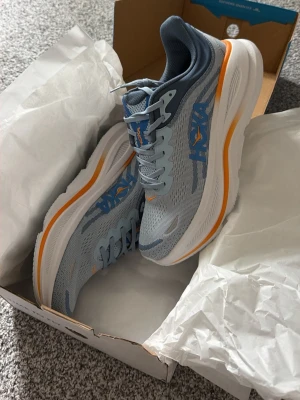 Hoka blå sneakers med orange detalj - Lätta sneakers från Hoka i ljusblå mesh med mörkblå detaljer och orange linje på den tjocka vita sulan. Skorna har Hoka-logga på sidan och är designade för maximal komfort och dämpning. Perfekta för dig som gillar sportig stil och vill ha något riktigt bekvämt.