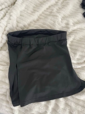 Svart skort med slits - Stilren svart topp utan band med hög midja och bred linning.  har en diskret slits på sidan och är sydd i ett mjukt, följsamt tyg som ger en snygg siluett. 