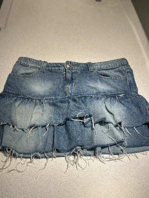 Blå jeanskjol med volanger - Trendig blå jeanskjol med två lager volanger och råa, fransiga kanter. Kjolen har klassiska fickor fram och bak samt hällor för skärp. Perfekt för dig som gillar en edgy och avslappnad stil.