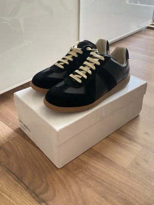 Svarta sneakers från Maison Margiela - Stilrena svarta sneakers från Maison Margiela med beigea skosnören och gummisula. Skorna har en klassisk låg siluett och är tillverkade i en mix av läder och mocka. Perfekta för dig som gillar minimalistisk och tidlös design.