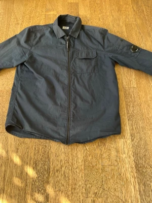 Mörkblå overshirt från C.P. Company - Mörkblå overshirt från C.P. Company. Köpte den personligen för nästan exakt två år sedan. Köpte den på NK Kids & Teens. Har använts väldigt mycket men är inte i för dåligt skick. 