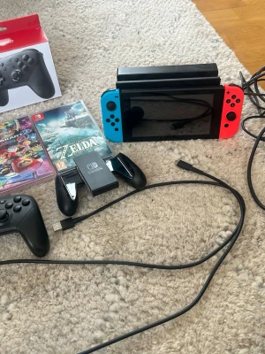 Nintendo Switch - Nintendo Switch med blå och röd Joy-Con, dockningsstation, Pro Controller, laddkabel och två spel: Mario Kart 8 Deluxe och The Legend of Zelda: Tears of the Kingdom. Konsolen är svart med färgglada handkontroller och tillbehör i plast. KNAPPT ANVÄND