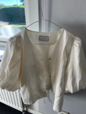 Krämvit blus med puffärm från Neo Noir - Stilren krämvit blus från Neo Noir med puffiga trekvartsärmar och v-ringning. Blusen har dekorativa knappar framtill och är tillverkad i ett lätt, vävt material som ger en luftig känsla. Perfekt för en trendig och avslappnad look.