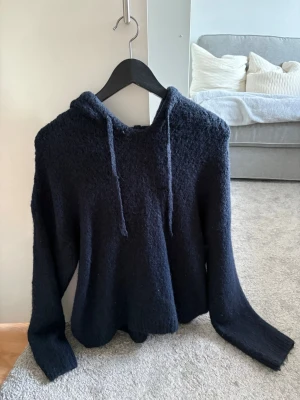 Mörkblå stickad hoodie med huva - Mysig mörkblå stickad hoodie med huva och dragsnören. Tröjan har en avslappnad passform och långa ärmar, perfekt för en chill och stilren look. Materialet är mjukt och fluffigt, vilket gör den extra bekväm att bära. 