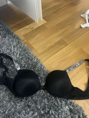 Svart push-up bh från Victoria's Secret - Säljer en svart push-up bh från Victoria's Secret med vadderade kupor och breda axelband med logotyptryck. Bh:n har släta kupor och är tillverkad i ett mjukt material som känns bekvämt mot huden. Perfekt för att ge extra lyft och stöd.