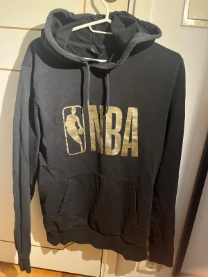 Svart NBA hoodie från New Era - Snygg svart hoodie från New Era med stort guldfärgat NBA-tryck på bröstet. Klassisk huva med snörning och en stor magficka framtill. Perfekt för dig som gillar basket och vill ha en stilren och sportig look.