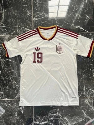 Vit Spanien fotbollströja Adidas - Säljer en vit retroinspirerad Spanien fotbollströja från Adidas med vinröda och gula detaljer. Tröjan har nummer 19 på bröstet, Adidas-logga och Spaniens landslagsemblem. Tillverkad i lätt och ventilerande material, perfekt för träning eller match.