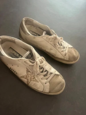 Golden Goose Super-Star sneakers - 💓Golden Goose Super-Star sneakers i vitt läder med en glittrig guldstjärna på sidan och silverdetalj på hälen. Skorna har vita snören, beige sula och en klassisk låg silhuett. Perfekta för dig som gillar en unik och trendig look. Använda så därav lågt skick. skick enligt bilder