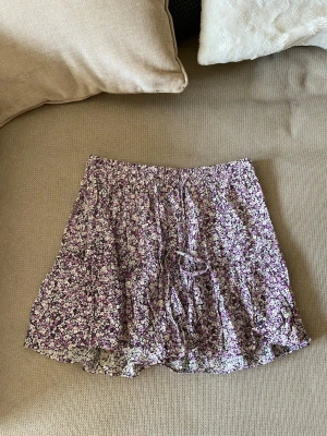 Lila blommig kjol från Zara - Supersöt kort kjol från Zara med lila och vita småblommor. Kjolen har resår i midjan och en liten knytning framtill. Den är luftig och har volanger nertill, perfekt för en trendig och avslappnad look. Kjolen har inbyggda shorts.