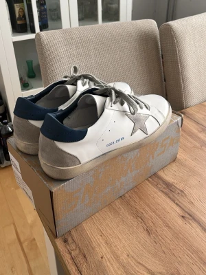 Golden Goose Superstar vita sneakers - Golden Goose Superstar sneakers i vitt läder med grå mockadetaljer och silverfärgad stjärna på sidan. Skorna har grå skosnören, blå hälkappa och klassisk låg siluett. Snygg och trendig modell med tydlig branding på plösen och insidan.