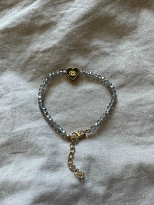 Armband med pärlor och hjärtdetalj - Snyggt armband med glittrande grå pärlor och en guldfärgad hjärtberlock med öga i mitten. Justerbar kedja i guldton ger en flexibel passform. Fint skick, hör av er om ni har frågor!