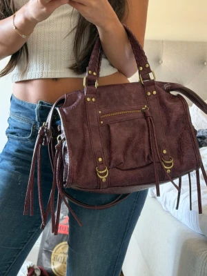 Vinröd handväska från Bershka - Snygg vinröd handväska i skinnimitation med flätade handtag och guldfärgade metalldetaljer. Väskan har en dragkedjeficka framtill och långa dekorativa remmar. Köpt för ett halvår sedan på Bershka men tyvärr inte riktigt kommit till användning så den är i superbra skick!
