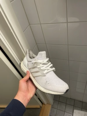 Vita Adidas Ultraboost sneakers - Säljer ett par vita Adidas Ultraboost sneakers med stickad mesh-överdel och klassiska tre ränder på sidan. Skorna har en tjock, responsiv Boost-sula och vita skosnören. Perfekta för dig som gillar stilrena och bekväma sneakers.