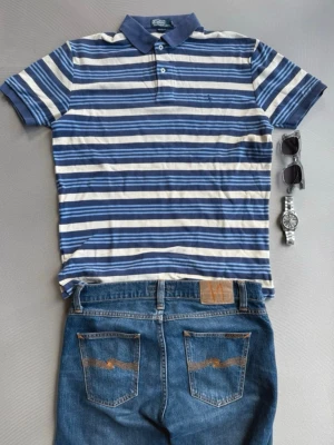 Randig pikétröja från Polo Ralph Lauren - Snygg kortärmad pikétröja från Polo Ralph Lauren med blå och vita horisontella ränder. Klassisk krage med knappar framtill och diskret logga på bröstet. Perfekt för en avslappnad och stilren look.
