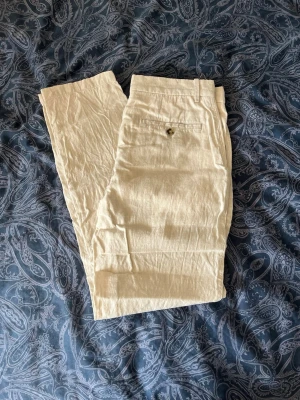 Beiga linnebyxor med rak passform - Säljer ett par stilrena beiga linnebyxor med rak passform och knappgylf. Byxorna har fickor fram och en bakficka med knapp. Perfekta för en avslappnad och trendig look under varma dagar. Materialet är svalt och luftigt linne.
