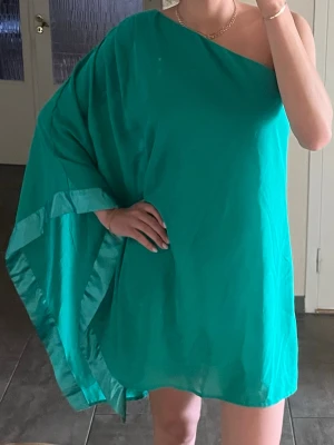 Grön oneshoulder kortklänning storlek M - Snygg grön/emeraldgrön kortklänning med oneshoulder-design och lös passform. Klänningen har ett brett ärm-parti på ena sidan och är ärmlös på andra. Storlek 38/M, men passar mig som vanligtvis bär S också pga den lösq passformen. 